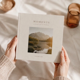 Hands holding a square cream photobook titled Moments, with a mountain landscape on the cover